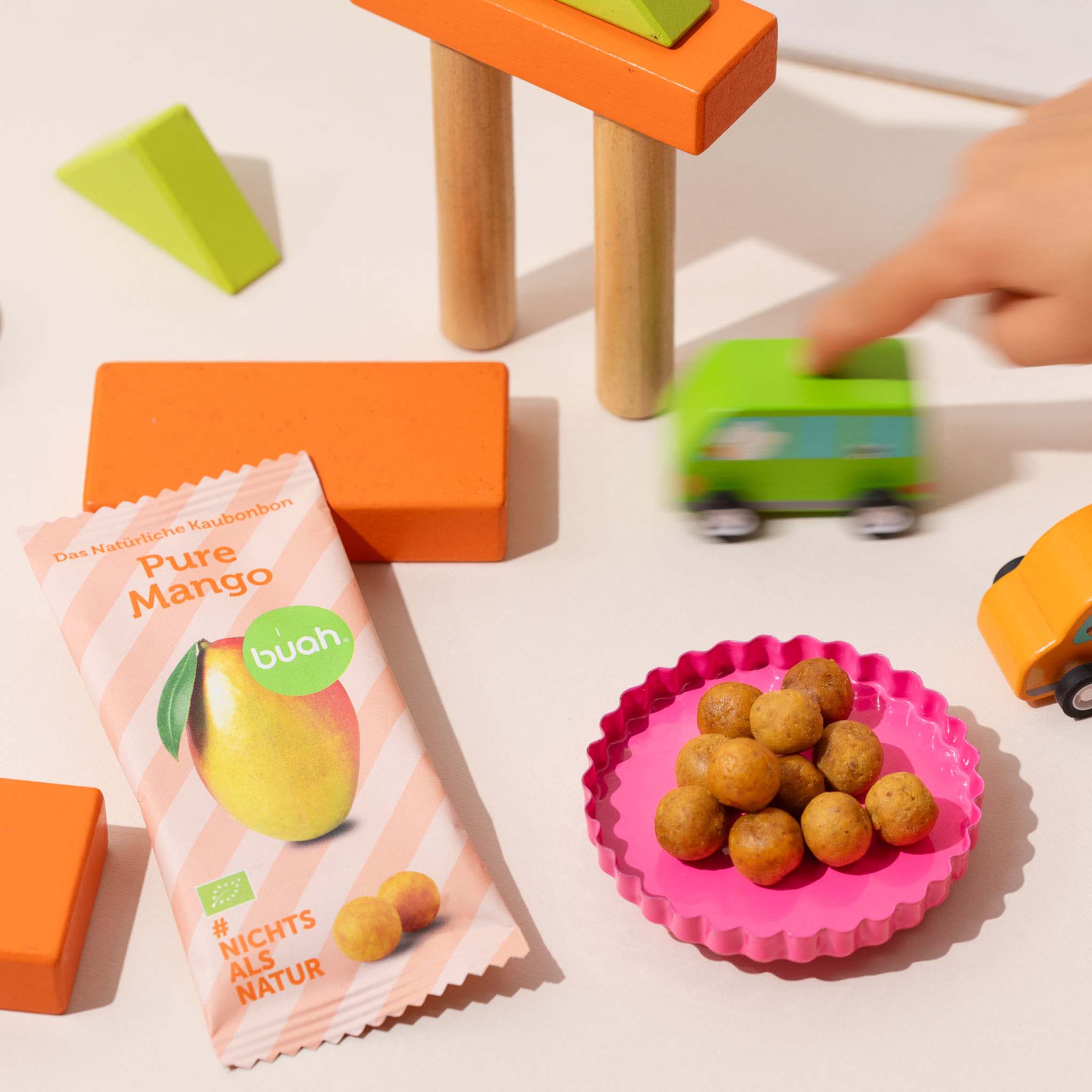 buah Kaubonbon Mango Packung neben Kaubonbons in Schale und Holzspielzeug im Hintergrund