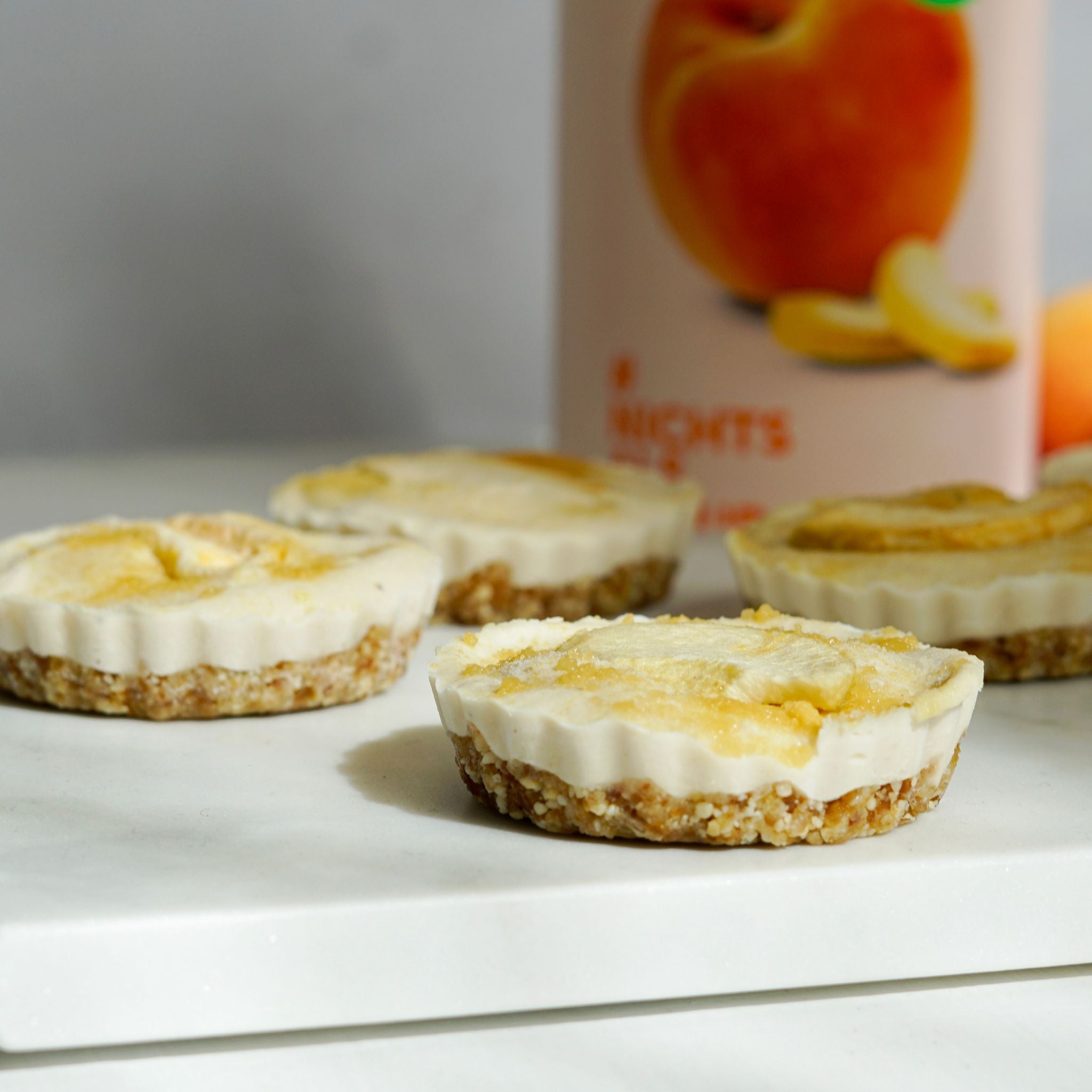Kleine flache Tartlets mit crunchy Base und Cremigem buah Pfirsich Topping, liegen schoen angerichtet nebeneinander, buah gefriergetrockneter Pfirsich in Dose im Hintergrund