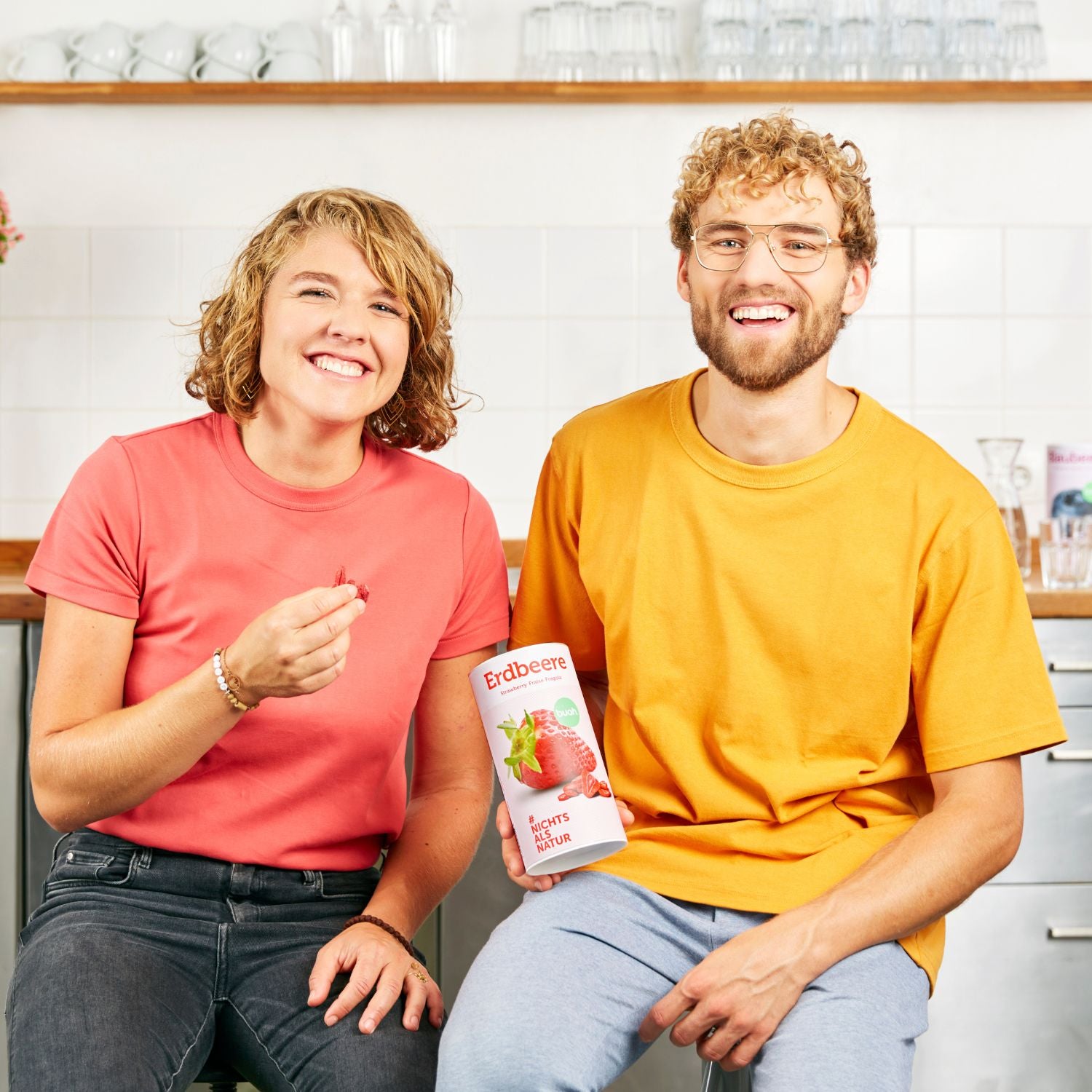 buah Gruender Jessica und Daniel sitzen nebeneinander mit einer Dose buah Erdbeeren und lachen in die Kamera