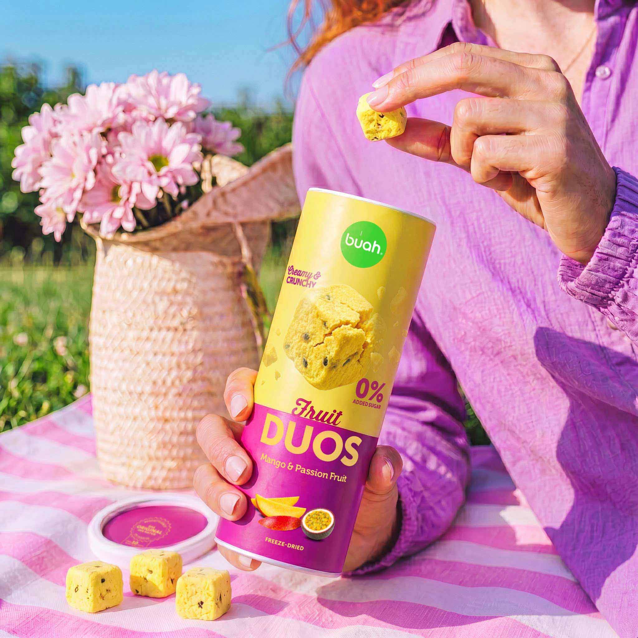 Eine Person sitzt auf einer Picknickdecke im Gras. Sie hält ein quadratisches Stück buah Fruit DUOS Mango & Maracuja zwischen Daumen und Zeigefinger. In der anderen Hand hält sie die geöffnete Slim-Dose, vor ihr liegen weitere Fruchtwürfel.