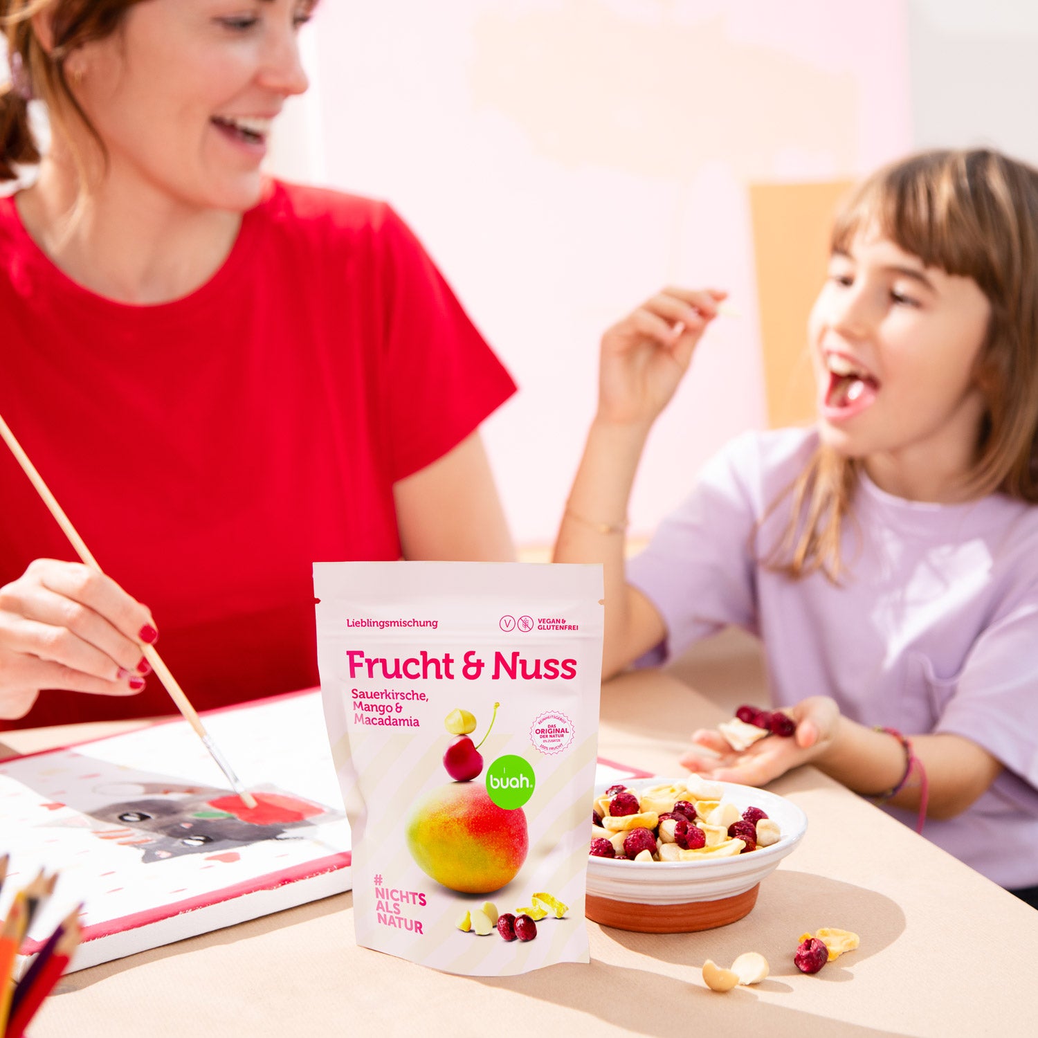 Neue buah Frucht & Nuss Mischung mit Macadamia steht auf einem Tisch, im Hintergrund ist eine Mama mit ihrer Tochter am Malen und Snacken