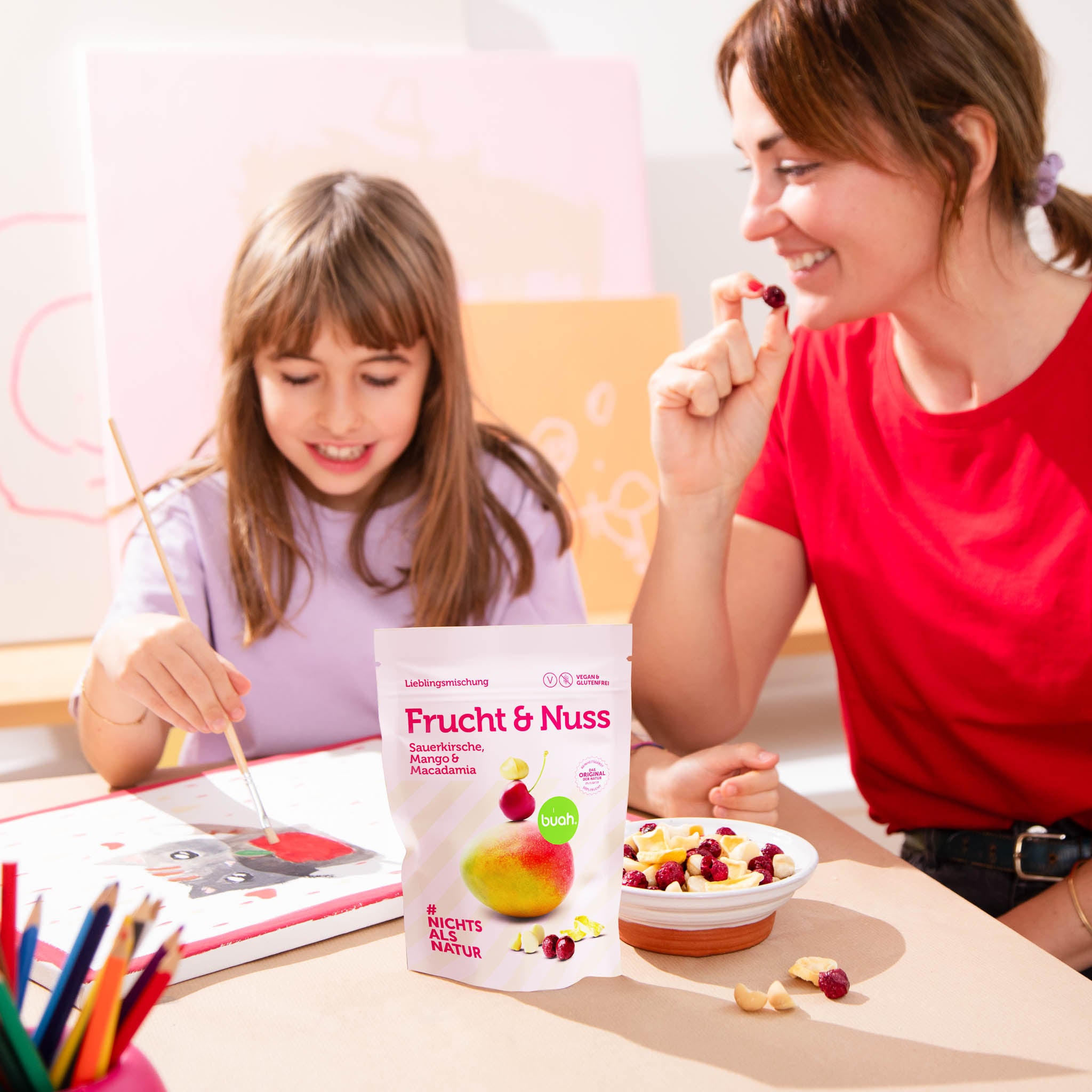 Neue buah Frucht & Nuss Mischung mit Macadamia steht auf einem Tisch, im Hintergrund ist eine Mama mit ihrer Tochter am Malen und Snacken