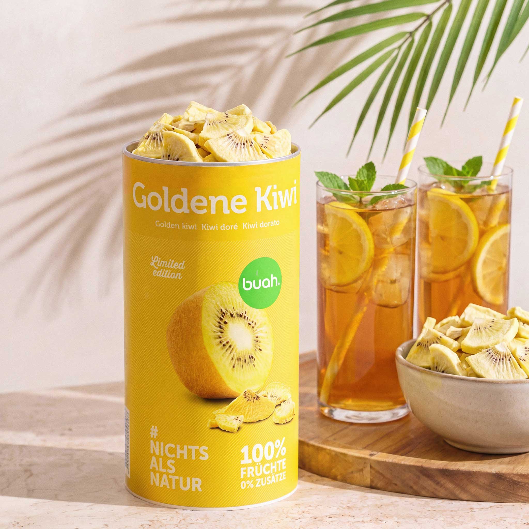 Ein erfrischendes Sommer-Arrangement. Eine geöffnete gelbe Dose buah gefriergetrocknete Goldene Kiwi steht neben zwei Gläsern Eistee mit Zitrone und Minze sowie einer Schale voller Kiwi-Stücke auf einem Holztablett. Palmblatt-Schatten fallen auf die Szene.