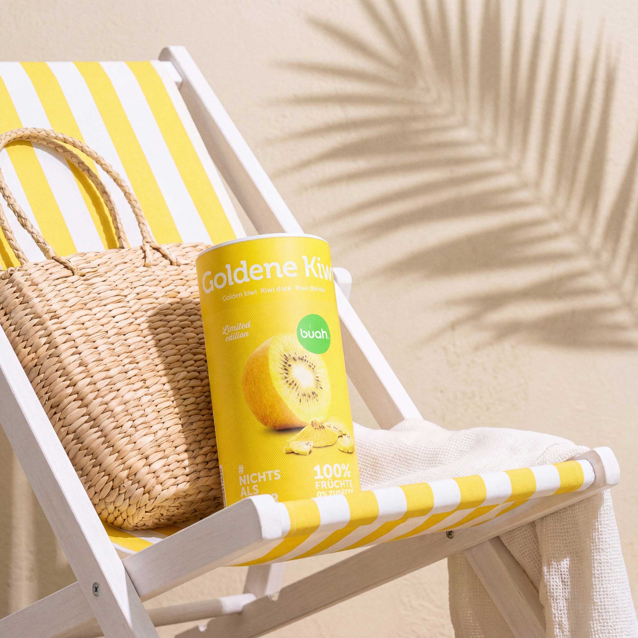 Eine gelbe Dose buah gefriergetrocknete Goldene Kiwi liegt in einem gelb-weiß gestreiften Liegestuhl. Daneben steht eine sommerliche Strohtasche, im Hintergrund zeichnet sich der Schatten von Palmblättern ab.