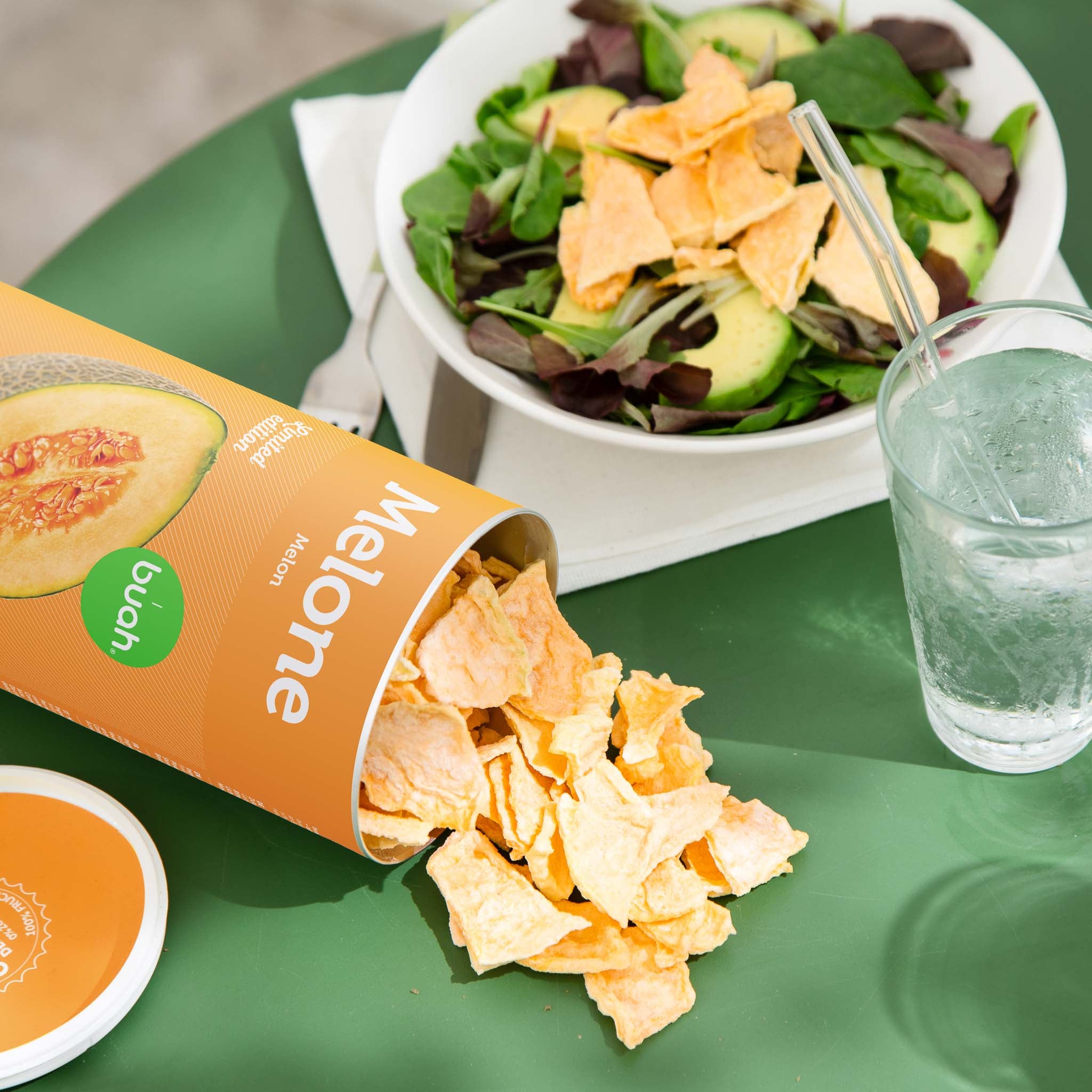 Auf gruenem Tisch liegt eine umgefallene buah Dose gefriergetrocknete Cantaloup Melone. Grosse knackige Stecke Melone liegen auf dem Tisch. 