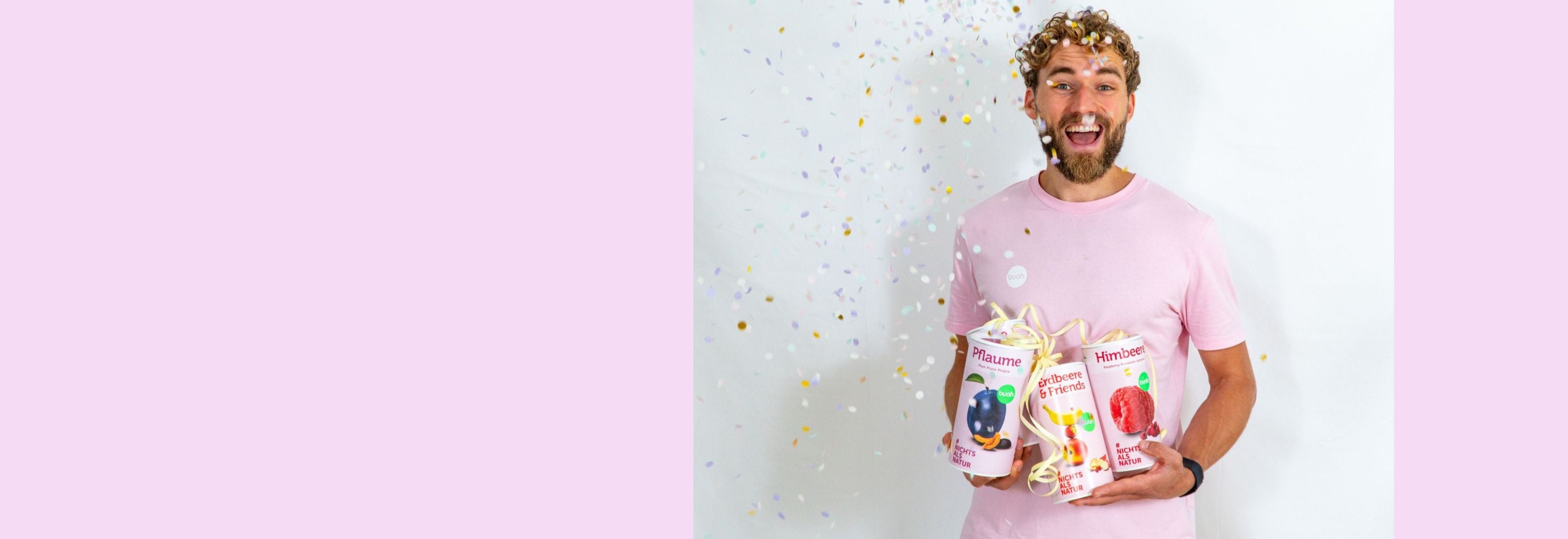 Daniel Gruender von buah in rosa buah Shirt mit buah gefriergetrockneten Fruechten in der Dose im Arm - Konfetti und Luftschlangen vor rosa Hintergrund