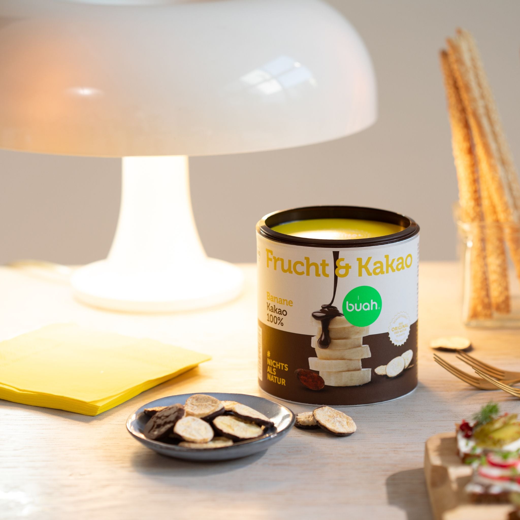 Kleine buah Dose Frucht & Kakao Banane als Schokofruechte steht auf Tisch neben Lampe, davor kleiner Teller mit gefriergetrockneter Banane, die mit Kakao ueberzogen ist