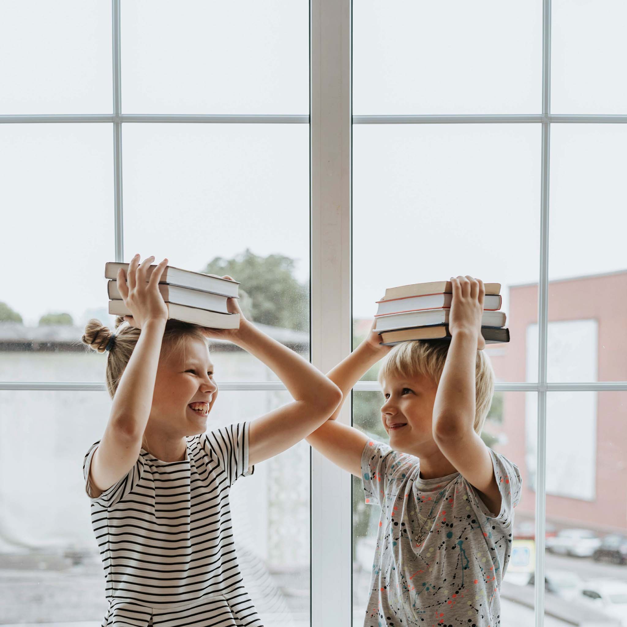Zwei lachende Schulkinder der Grundschule halten Buecher gestapelt auf dem Kopf