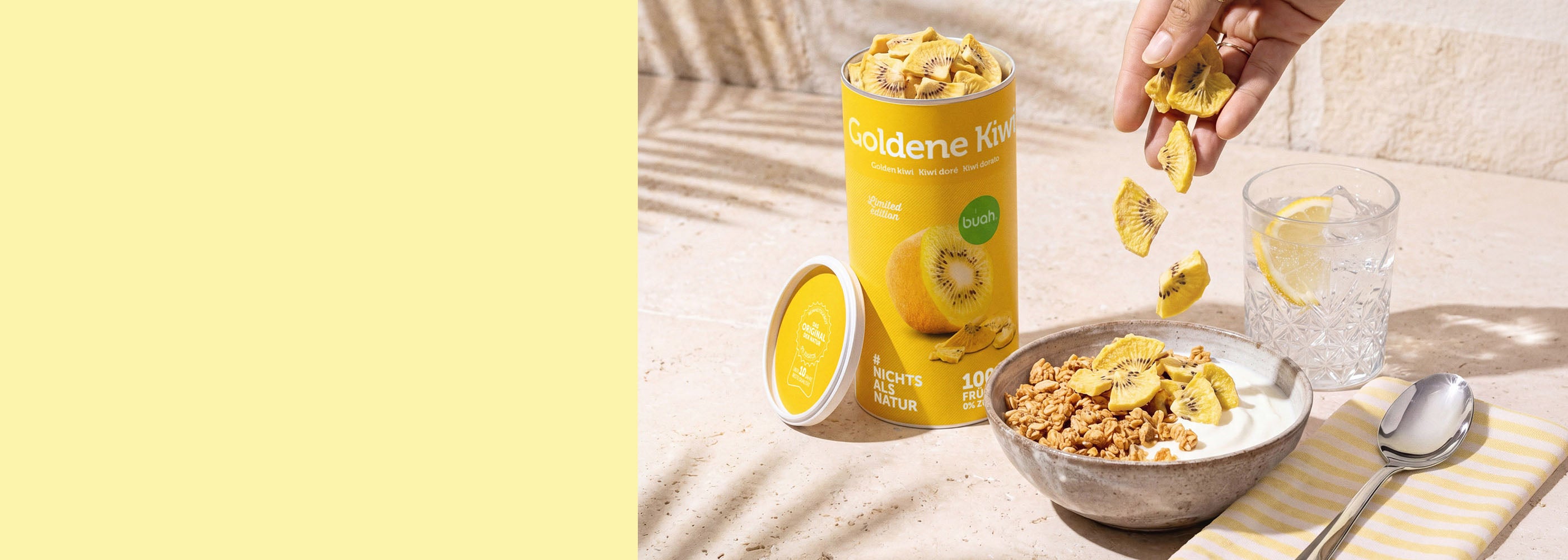 Ein breites Banner. Auf der rechten Seite stehen eine gelbe Dose buah gefriergetrocknete Goldene Kiwi und eine Schale Müsli mit Joghurt auf einer hellen Steinplatte. Eine Hand streut Kiwi-Stücke darüber. Daneben steht ein Glas Wasser mit Zitrone. Die linke Bildhälfte besteht aus einer hellgelben Freifläche.