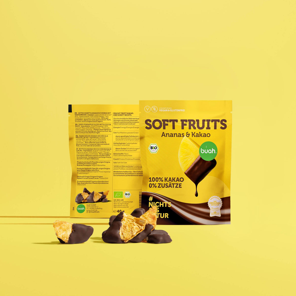 Zwei buah SOFT FRUITS Snackpacks Ananas & Kakao vor gelbem Hintergrund. Davor liegen mehrere Ananasscheiben, teils mit dunkler Schokolade überzogen. Auf der Vorderseite steht „100% KAKAO 0% ZUSÄTZE“ und „#NICHTS ALS NATUR“.