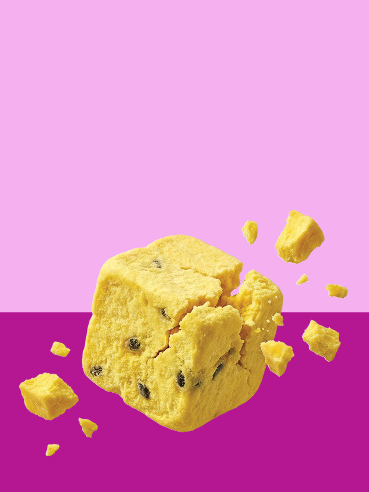 Ein zerbrechender buah gefriergetrockneter Fruchtwürfel der Sorte Mango & Maracuja schwebt im Hochformat. Die obere Bildhälfte ist hellrosa, die untere dunkelpink. Kleine gelbe Stückchen fliegen um den Würfel.